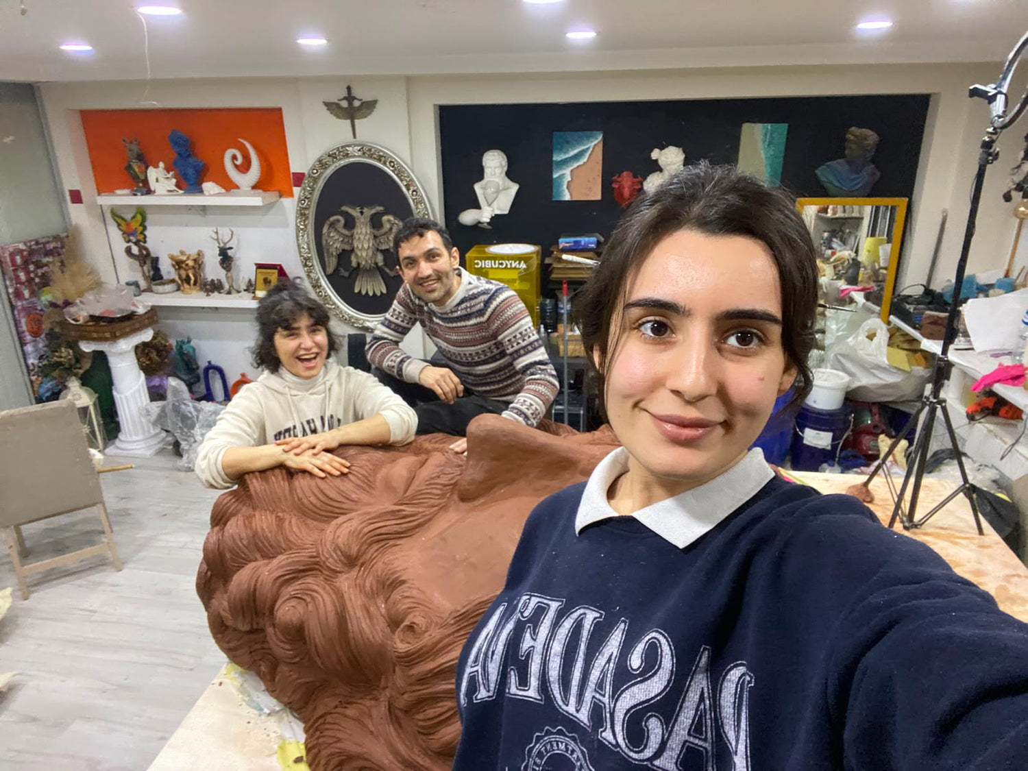 Atölyede üç kişi, büyük kahverengi kabartmalı heykel çalışmasıyla birlikte