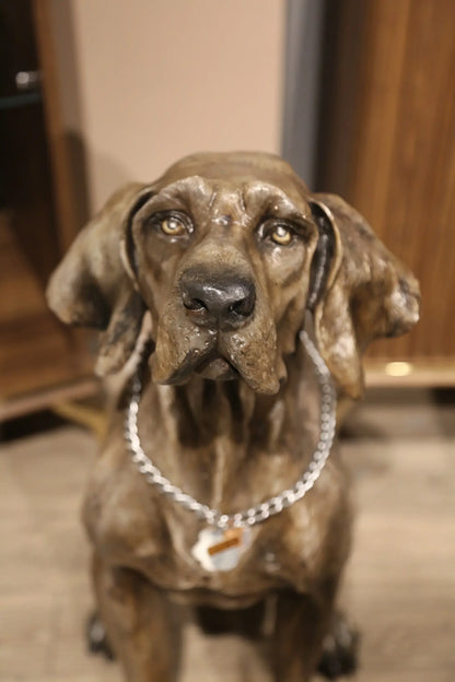 Metalik renkli av köpeği heykeli, boynunda zincir tasma ile iç mekanda sergileniyor.